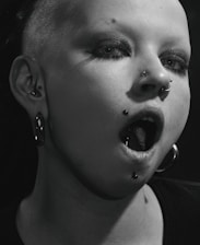 A close-up of a person with heavy makeup and multiple piercings, including a nose ring and lip ring. Their hair is shaved on one side, and they are wearing large hoop earrings. The lighting casts a dramatic shadow across their face, enhancing the edgy aesthetic.