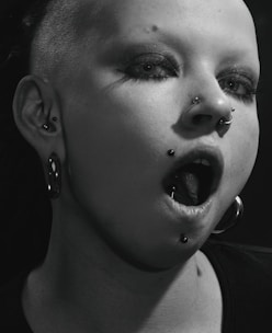 A close-up of a person with heavy makeup and multiple piercings, including a nose ring and lip ring. Their hair is shaved on one side, and they are wearing large hoop earrings. The lighting casts a dramatic shadow across their face, enhancing the edgy aesthetic.