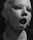 A close-up of a person with heavy makeup and multiple piercings, including a nose ring and lip ring. Their hair is shaved on one side, and they are wearing large hoop earrings. The lighting casts a dramatic shadow across their face, enhancing the edgy aesthetic.