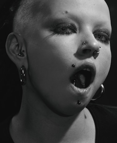 A close-up of a person with heavy makeup and multiple piercings, including a nose ring and lip ring. Their hair is shaved on one side, and they are wearing large hoop earrings. The lighting casts a dramatic shadow across their face, enhancing the edgy aesthetic.