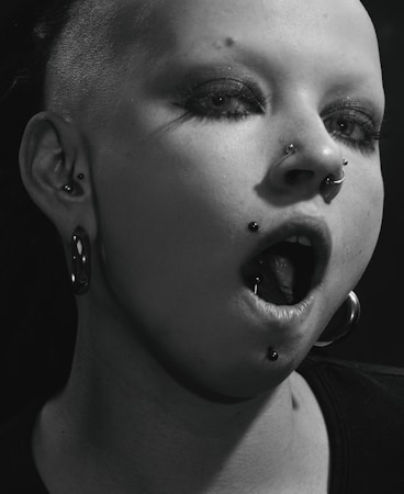 A close-up of a person with heavy makeup and multiple piercings, including a nose ring and lip ring. Their hair is shaved on one side, and they are wearing large hoop earrings. The lighting casts a dramatic shadow across their face, enhancing the edgy aesthetic.