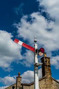 A traditional railway semaphore signal stands against a vibrant blue sky with fluffy white clouds. The signal's red and white arm is positioned horizontally, indicating a stop or caution message. In the background, part of a stone building with chimney stacks is visible, adding a rustic charm to the scene.