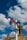 A traditional railway semaphore signal stands against a vibrant blue sky with fluffy white clouds. The signal's red and white arm is positioned horizontally, indicating a stop or caution message. In the background, part of a stone building with chimney stacks is visible, adding a rustic charm to the scene.