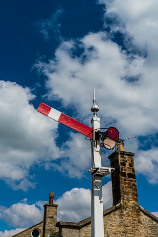 A traditional railway semaphore signal stands against a vibrant blue sky with fluffy white clouds. The signal's red and white arm is positioned horizontally, indicating a stop or caution message. In the background, part of a stone building with chimney stacks is visible, adding a rustic charm to the scene.