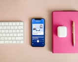 A neatly arranged workspace features a Bluetooth keyboard, a smartphone displaying a podcast app with an episode titled 'DeFi: Is Trustless More Trustworthy?', a pair of wireless earbuds in their case, a pink notebook, and a gold pen on a beige surface.
