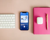 A neatly arranged workspace features a Bluetooth keyboard, a smartphone displaying a podcast app with an episode titled 'DeFi: Is Trustless More Trustworthy?', a pair of wireless earbuds in their case, a pink notebook, and a gold pen on a beige surface.