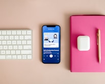 A neatly arranged workspace features a Bluetooth keyboard, a smartphone displaying a podcast app with an episode titled 'DeFi: Is Trustless More Trustworthy?', a pair of wireless earbuds in their case, a pink notebook, and a gold pen on a beige surface.