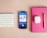 A neatly arranged workspace features a Bluetooth keyboard, a smartphone displaying a podcast app with an episode titled 'DeFi: Is Trustless More Trustworthy?', a pair of wireless earbuds in their case, a pink notebook, and a gold pen on a beige surface.
