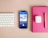 A neatly arranged workspace features a Bluetooth keyboard, a smartphone displaying a podcast app with an episode titled 'DeFi: Is Trustless More Trustworthy?', a pair of wireless earbuds in their case, a pink notebook, and a gold pen on a beige surface.