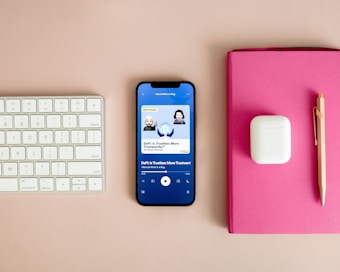 A neatly arranged workspace features a Bluetooth keyboard, a smartphone displaying a podcast app with an episode titled 'DeFi: Is Trustless More Trustworthy?', a pair of wireless earbuds in their case, a pink notebook, and a gold pen on a beige surface.