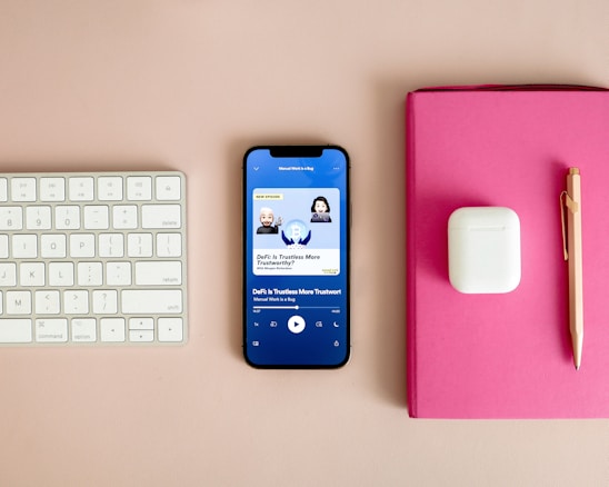 A neatly arranged workspace features a Bluetooth keyboard, a smartphone displaying a podcast app with an episode titled 'DeFi: Is Trustless More Trustworthy?', a pair of wireless earbuds in their case, a pink notebook, and a gold pen on a beige surface.