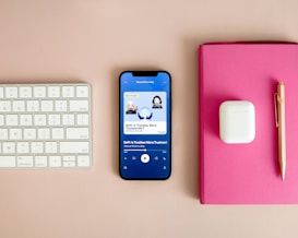 A neatly arranged workspace features a Bluetooth keyboard, a smartphone displaying a podcast app with an episode titled 'DeFi: Is Trustless More Trustworthy?', a pair of wireless earbuds in their case, a pink notebook, and a gold pen on a beige surface.