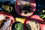 A variety of Mexican dishes with grilled meat and tortillas are placed on pink plates. A selection of sides including green chili peppers, lime wedges, and green salsa are served in small black bowls.