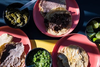 A variety of Mexican dishes with grilled meat and tortillas are placed on pink plates. A selection of sides including green chili peppers, lime wedges, and green salsa are served in small black bowls.