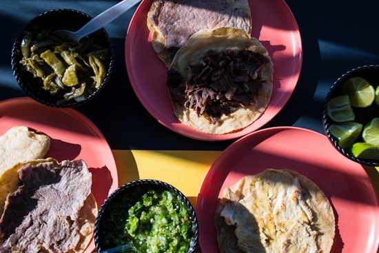 A variety of Mexican dishes with grilled meat and tortillas are placed on pink plates. A selection of sides including green chili peppers, lime wedges, and green salsa are served in small black bowls.