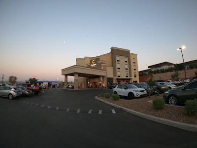 A modern multi-story hotel building set against a dusk sky, featuring a clearly visible sign for Comfort Suites. The foreground shows a parking lot containing several cars, with well-maintained landscaping and a few trees. A gas station can be seen in the background on the left side.