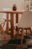 A wooden dining table with a decorative vase on top, surrounded by modern white chairs. The setting appears to be an indoor space with a patterned rug underneath the table.