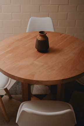 A stylish dining nook with a round wooden table, woven chairs, and a vase of dried flowers against a warm-toned wall.