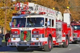 A red and white fire truck from the Siler City Fire Department is prominently displayed, adorned with a festive Christmas wreath on the front. A firefighter in uniform is situated on top, while other crew members are visible inside and around the vehicle. The background shows autumn-colored trees and additional emergency vehicles, with several people observing the scene.