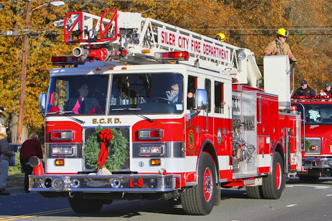 A red and white fire truck from the Siler City Fire Department is prominently displayed, adorned with a festive Christmas wreath on the front. A firefighter in uniform is situated on top, while other crew members are visible inside and around the vehicle. The background shows autumn-colored trees and additional emergency vehicles, with several people observing the scene.
