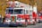 A red and white fire truck from the Siler City Fire Department is prominently displayed, adorned with a festive Christmas wreath on the front. A firefighter in uniform is situated on top, while other crew members are visible inside and around the vehicle. The background shows autumn-colored trees and additional emergency vehicles, with several people observing the scene.