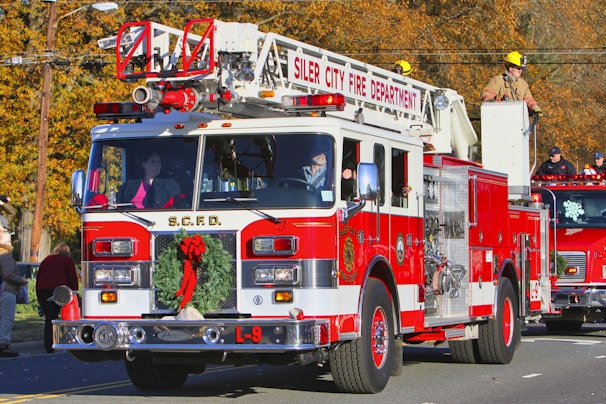 A red and white fire truck from the Siler City Fire Department is prominently displayed, adorned with a festive Christmas wreath on the front. A firefighter in uniform is situated on top, while other crew members are visible inside and around the vehicle. The background shows autumn-colored trees and additional emergency vehicles, with several people observing the scene.