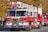 A red and white fire truck from the Siler City Fire Department is prominently displayed, adorned with a festive Christmas wreath on the front. A firefighter in uniform is situated on top, while other crew members are visible inside and around the vehicle. The background shows autumn-colored trees and additional emergency vehicles, with several people observing the scene.