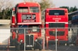 A vintage double-decker red bus is parked next to a modern red bus behind a chain-link fence. The double-decker bus displays various destinations and appears worn, while the modern bus shows the name Unitrans. Both buses are positioned in a parking area with some trees and buildings visible in the background.