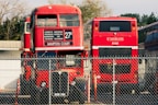 A vintage double-decker red bus is parked next to a modern red bus behind a chain-link fence. The double-decker bus displays various destinations and appears worn, while the modern bus shows the name Unitrans. Both buses are positioned in a parking area with some trees and buildings visible in the background.