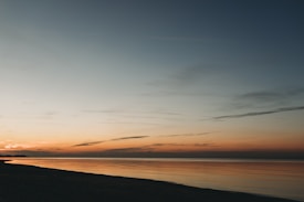 A serene horizon with a soft gradient transitioning from warm orange to cool blue, reflecting on a calm sea. Silhouettes of distant landform can be seen to the left, enhancing the tranquility of the scene.