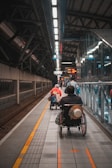 A wheelchair user is navigating through a subway platform, surrounded by the architectural elements of the station. The scene includes overhead lights, signs, and tracks, with a sense of movement as people travel along the platform.