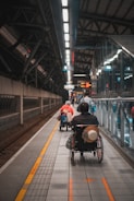 A wheelchair user is navigating through a subway platform, surrounded by the architectural elements of the station. The scene includes overhead lights, signs, and tracks, with a sense of movement as people travel along the platform.