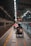A wheelchair user is navigating through a subway platform, surrounded by the architectural elements of the station. The scene includes overhead lights, signs, and tracks, with a sense of movement as people travel along the platform.