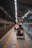 A wheelchair user is navigating through a subway platform, surrounded by the architectural elements of the station. The scene includes overhead lights, signs, and tracks, with a sense of movement as people travel along the platform.