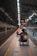 A wheelchair user is navigating through a subway platform, surrounded by the architectural elements of the station. The scene includes overhead lights, signs, and tracks, with a sense of movement as people travel along the platform.