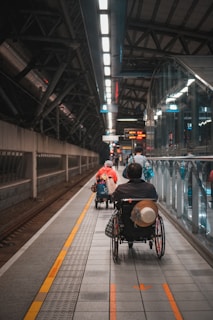 A wheelchair user is navigating through a subway platform, surrounded by the architectural elements of the station. The scene includes overhead lights, signs, and tracks, with a sense of movement as people travel along the platform.