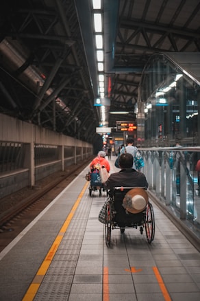A wheelchair user is navigating through a subway platform, surrounded by the architectural elements of the station. The scene includes overhead lights, signs, and tracks, with a sense of movement as people travel along the platform.