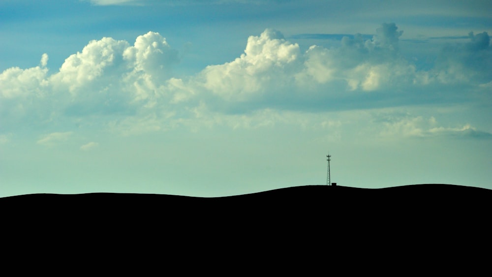 silueta de una persona parada en una colina bajo nubes blancas y un cielo azul durante el día