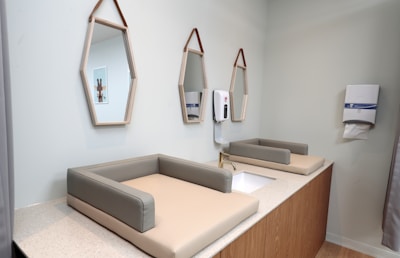 A modern and clean baby changing station is set in a softly lit room. The station includes two padded changing tables with a faucet and sink in between. Above the sink, two geometric mirrors hang with leather straps. A hand sanitizer dispenser and a paper towel holder are mounted on the wall. Soft pastel colors and wooden elements create a cozy atmosphere.