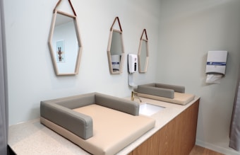 A modern and clean baby changing station is set in a softly lit room. The station includes two padded changing tables with a faucet and sink in between. Above the sink, two geometric mirrors hang with leather straps. A hand sanitizer dispenser and a paper towel holder are mounted on the wall. Soft pastel colors and wooden elements create a cozy atmosphere.