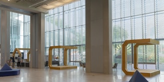 A spacious interior with large floor-to-ceiling windows allowing natural light to illuminate the open area. The modern architectural design includes geometric structures and minimalist furnishings. Two people are seen near a desk or kiosk on the left. The walls and ceilings are made of concrete with metal accents, and several triangular seating areas are scattered throughout the space.