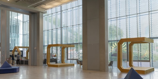 A spacious interior with large floor-to-ceiling windows allowing natural light to illuminate the open area. The modern architectural design includes geometric structures and minimalist furnishings. Two people are seen near a desk or kiosk on the left. The walls and ceilings are made of concrete with metal accents, and several triangular seating areas are scattered throughout the space.