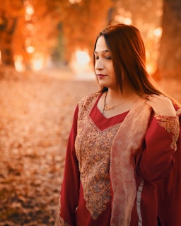 A woman in traditional attire stands in an autumnal setting. Her outfit is a deep red with intricate gold embroidery. She gently holds her shawl while looking slightly downward, with her hair cascading over her shoulders. The background is filled with fallen leaves and trees in shades of orange and gold, suggesting a warm, serene setting.