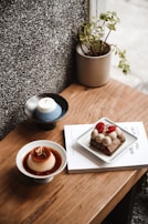 A cozy arrangement on a wooden table includes a small potted plant, a dessert with caramel sauce in a white bowl, a slice of cake garnished with raspberries and cream on a square plate, a candle on a dark dish, and a book partially visible beneath the plate.