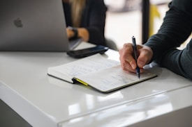 A person is writing in a notebook on a white desk, with a laptop open nearby. Another individual is partially visible in the background. A smartphone is placed on the desk beside the notebook.