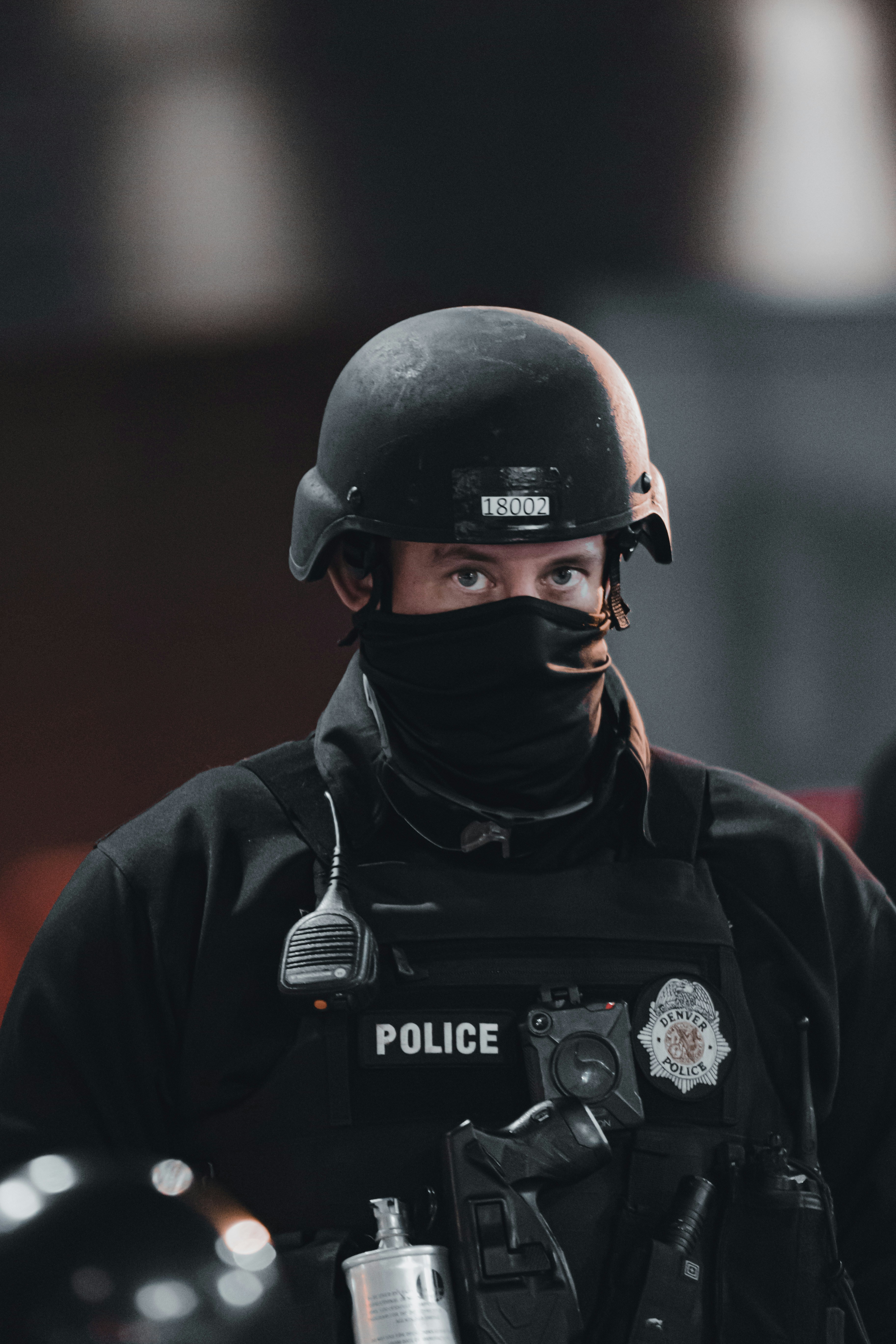 A police officer wearing tactical gear, including a black helmet and a face cover, is seen. The officer's uniform displays a police badge and has various equipment attached, including a radio and other tactical gear.