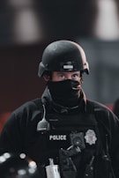 A police officer wearing tactical gear, including a black helmet and a face cover, is seen. The officer's uniform displays a police badge and has various equipment attached, including a radio and other tactical gear.