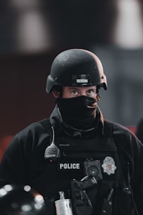 A police officer wearing tactical gear, including a black helmet and a face cover, is seen. The officer's uniform displays a police badge and has various equipment attached, including a radio and other tactical gear.