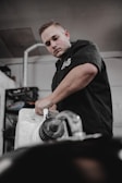 A person in a black shirt is working with a large plastic container, possibly a gasoline or liquid container, inside a workshop or garage. The setting includes industrial elements like metal pipes and shelves in the background.