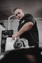A person in a black shirt is working with a large plastic container, possibly a gasoline or liquid container, inside a workshop or garage. The setting includes industrial elements like metal pipes and shelves in the background.